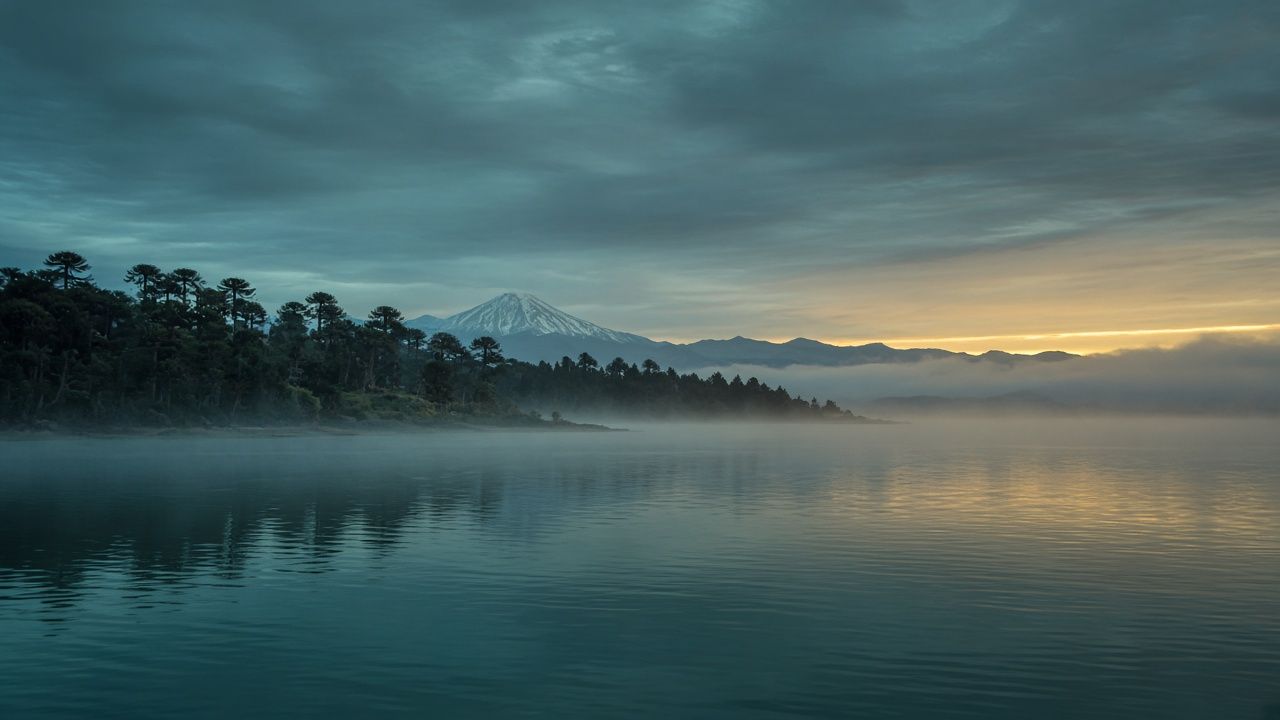 Amanecer sobre el lago con niebla, orilla de araucarias y volcán en la distancia bajo una franja dorada de luz